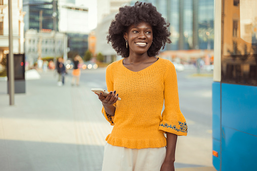 Cheerful African Woman Holds Smartphone and Looking Away. Smiles and Walks Outdoors
