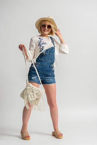 Happy young blonde woman in rustic casual clothes, sunglasses and a hat with a handbag in her hand posing on a white background