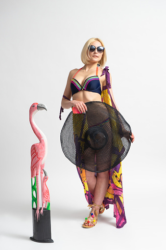 Young blonde woman in a swimsuit, sandals, sunglasses and a pareo with a black beach hat in her hand posing next to a flamingo figurine on a white background