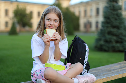 A cheerful school girl eating sandwich on a school park bench. She is a cute pupil having a break from her studies with her backpack and trees.