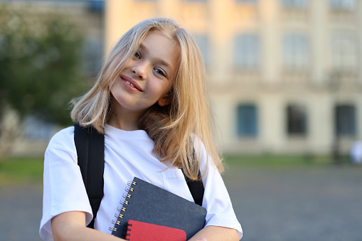 A schoolgirl with a backpack holding a notebook in front of a building. She is a cute pupil going to school and reading her book.