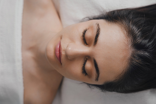 Top view of young ethnic female with closed eyes lying on bed and touching face while resting at home