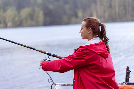 Under a red raincoat, a young woman enjoys fishing on the serene lake. With a fishing rod in hand and a metal water bottle nearby, she relishes the peaceful moment amid nature's embrace.