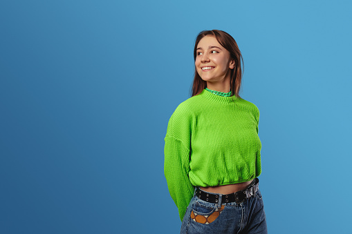 Cheerful young lady wearing green sweater, smiling and looking at copy space while standing against blue background
