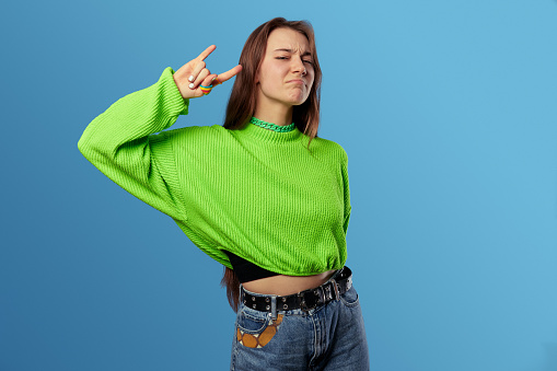 Beautiful young girl wearing casual sweater doing rock symbol with hand up while looking at camera, isolated over blue background.