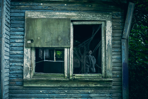 A creepy doll covered in cobwebs stands in the window of an abandoned house.