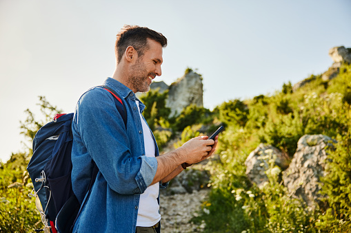 Smiling adult man hiking in mountains with backpack navigating with mobile phone