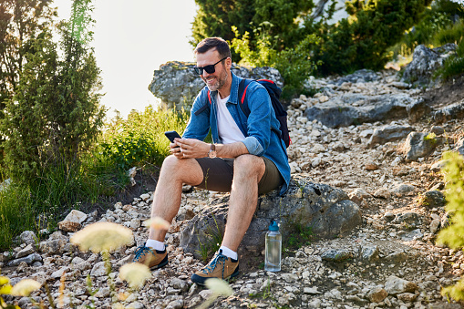 Happy adult man sitting on rock using his phone while hiking