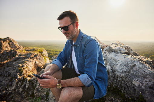 Adult man using his phone sitting on the rock while hiking in Jura, Poland