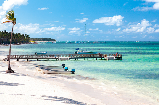 A wooden pier extends into the clear blue-green ocean at a tropical beach. The pier has a boat docked at the end and there are more boats in the distance. The beach has white sand and palm trees. The sky is blue with some clouds. A small group of people are on the pier, enjoying the view. This image can be used for social, leisure, or adventure themes.