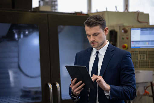 An engineer in a blue suit is checking data on a wifi tablet and talking to an Asian worker in a uniform in production.