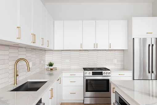 A kitchen detail with white cabinets, gold hardware and faucet, marble countertops, and a brown picket tile backsplash.