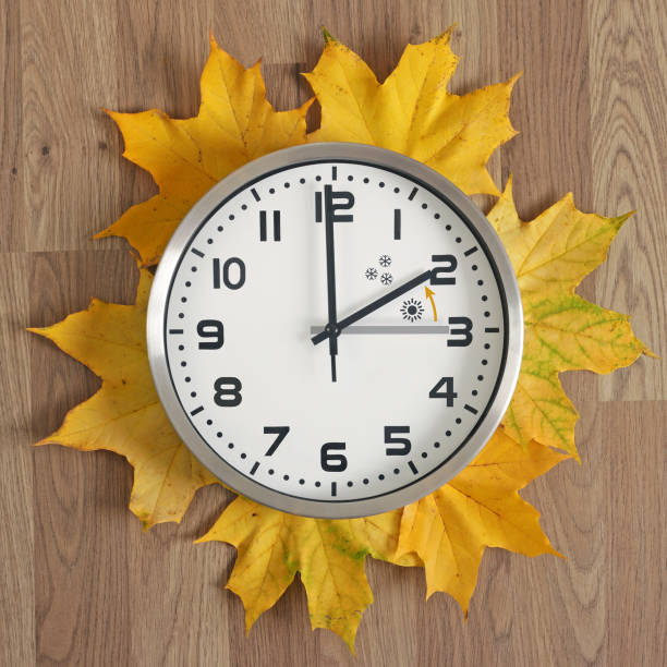 une horloge, l’aiguille grise pointe à trois heures, la noire une à deux heures.   des feuilles d’automne tombées jaunes traînent. déplacer les mains vers l’arrière. - minute hand number 15 clock time photos et images de collection