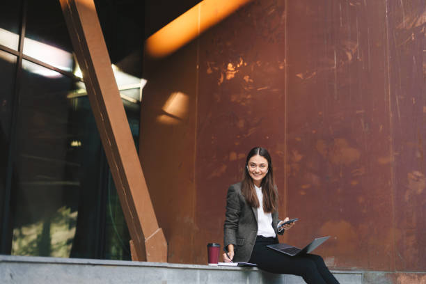 istock Attractive brunette woman sitting on the stairs and working on laptop while using mobile phone. Positive human face expression, emotion, success concept 1587602083