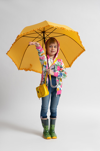Little girl in casual autumn clothes and rubber boots pretends to hide from the rain under a yellow umbrella on a white background