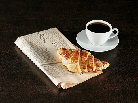 cup of coffee with milk foam, croissant, orange juice,    stands on a table in cafeteria with newspaper