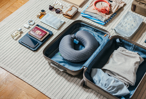 Close up shot of an open suitcase with various stuff laid on the floor ready for a trip away. There is passport, mobile phone, tablet, perfume, sunglasses, wallet, headphones, charger, clothes and a travel pillow. There is no one on the photo.