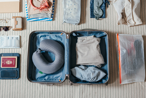 Shot from above of the items laid on the floor. Someone is preparing for a trip and packing a suitcase. There are sunglasses, wallet, clothes, travel pillow, open suitcase, passport, mobile phone, and other essentials for a holiday.