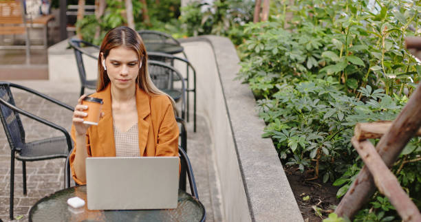 Caucasian businesswoman sitting at work wearing headphones Online meeting via laptop alone in the park. Woman drinking coffee alone in summer cafe stock photo