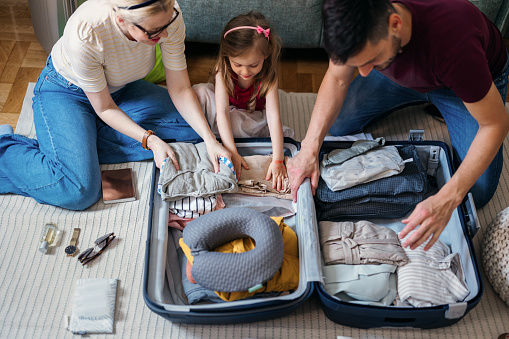 A couple with the little girl smiling and getting ready for travelling. They are sitting on the floor with various essential  stuff around them and packing them into the suitcase. A little girl is sitting in between and helping them put things in.