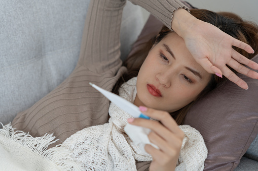 Young Asian woman having high fever while checking body temperature, female sneezing and runny nose with seasonal influenza, allergic, digital thermometer, virus, coronavirus, illness, respiratory