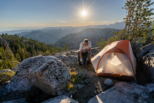 Mature Caucasian man, hiker, backpacker, sitting on a rock by his tent preparing dinner as the sun goes down in the distance, Seneca Lake, Wind River Range, Wyoming