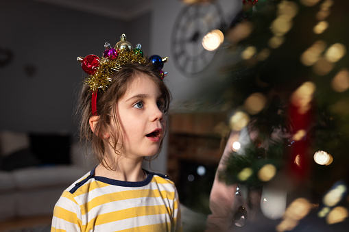 A young girl in awe looking at a Christmas tree which has string lights on it illuminating her face. She has baubles and tinsel on a headband over her head.