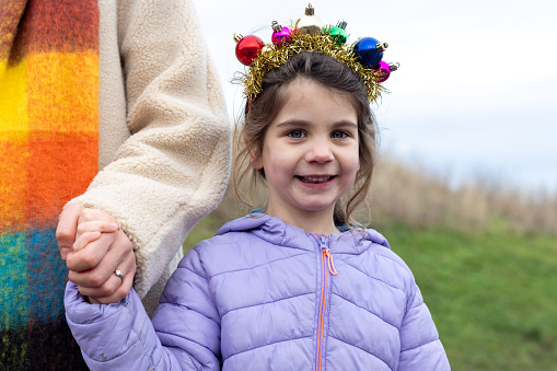 Waist-up shot of a young girl looking into the camera. She is wearing a warm jacket and holding hands with her unrecognisable mother. She has a Christmas costume item on her head.