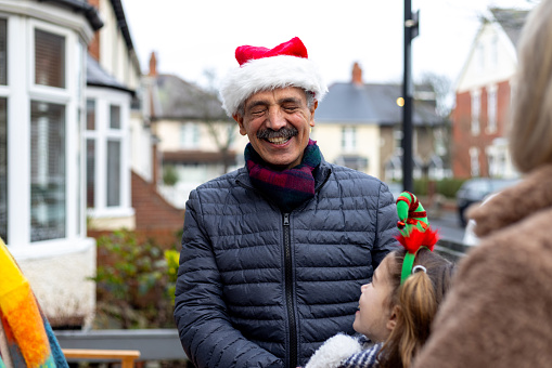 Over-the-shoulder shot of a grandfather laughing with his family standing outside their family home. He is wearing a warm jacket and a Santa hat.