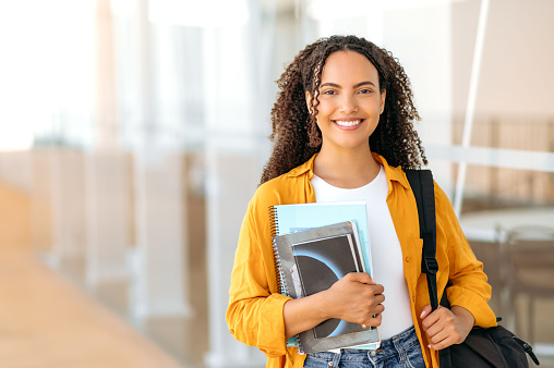 Happy lovely curly haired brazilian or hispanic female student, with a backpack, hold books and notebooks in her hand, stand near the university campus, looks and smile at camera. Copy-space