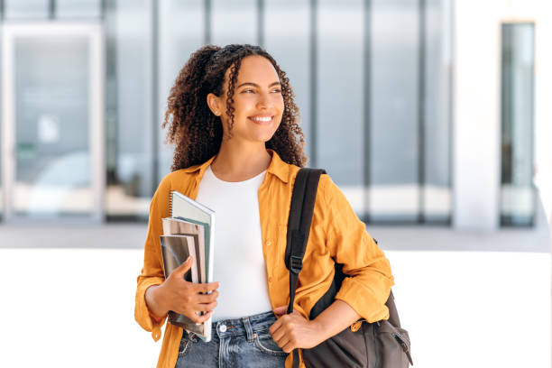 feliz estudiante. estudiante positiva, de nacionalidad brasileña o hispana, con una mochila, sosteniendo libros y cuadernos en la mano, parada cerca del campus universitario, mira hacia otro lado y sonríe - alumnos fotografías e imágenes de stock