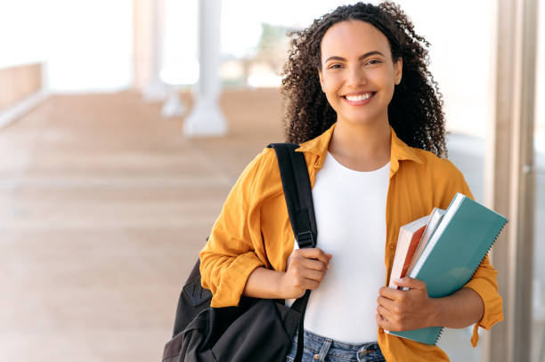 Close-up of a cheerful lovely curly haired brazilian or hispanic female student, with a backpack, hold books and notebooks in her hand, stand near the university campus, looks and smile at camera stock photo