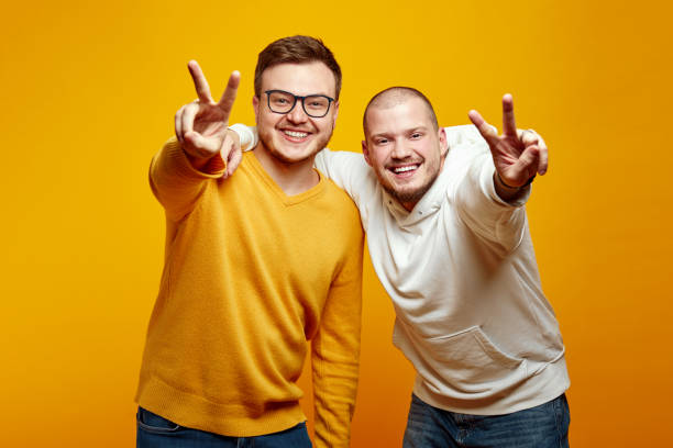 istock Two attractive excited young men embracing each other and showing peace gesture against yellow wall 1587760871