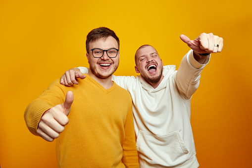 Portrait of two handsome happy young men embracing each other and showing thumbs up while laughing at camera, standing isolated over yellow orange studio background. True friendship concept