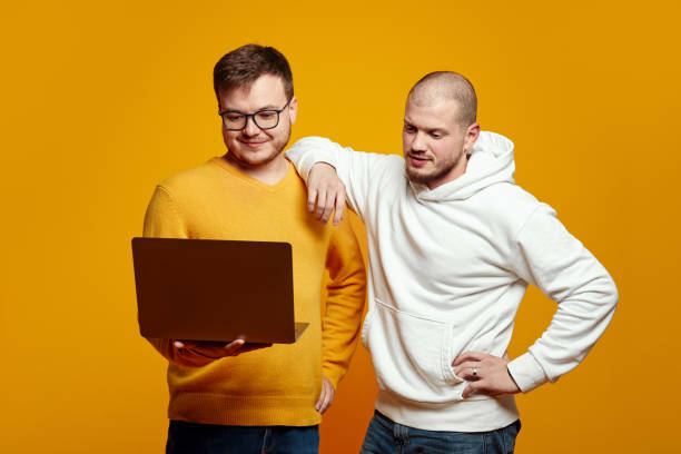 istock Two handsome young men holding and using laptop, looking at notebook, isolated over yellow background 1587771060