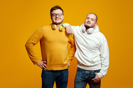 Two young men with headphones wearing casual clothes, smiling and looking at camera while standing isolated over yellow background. True friendship concept