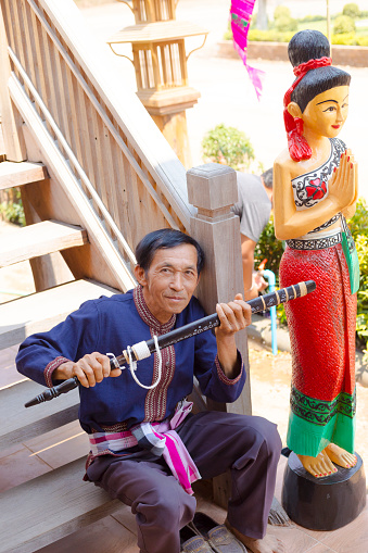 Mature thai man is sitting on steps and is showing old sword. Man is siting on steps of cultural house and center in On Tai, Chiang Mai, San Kamphaeng district of Chiang Mai. Man is dressed in traditional clothing. At his left side is a wooden female thai woman statue in traditional clothings welcoming people with Wai gesture