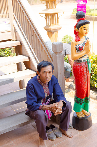 Mature thai man is sitting relaxed on steps of old wooden house. Man is siting on steps of cultural house and center in On Tai, Chiang Mai, San Kamphaeng district of Chiang Mai. Man is dressed in traditional clothing. At his left side is a wooden female thai woman statue in traditional clothings welcoming people with Wai gesture
