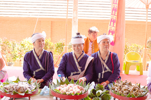 Group of blue dressed mature Thai women with traditional clothes. Women are  sitting behind  local thai food. Women are sitting on floor and are wearing headscarf. Scene is at cultural center house in On Tai, Chiang Mai, San Kamphaeng district of Chiang Mai. District is center os traditional northern thai art and craft and textile industry