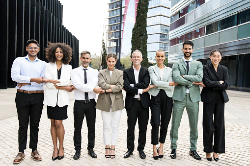 Group of smiling multiethnic businessman and businesswoman holding hands staring at camera outside. Multiracial cheerful confident executive team standing in a line in the street. Teamwork concept.