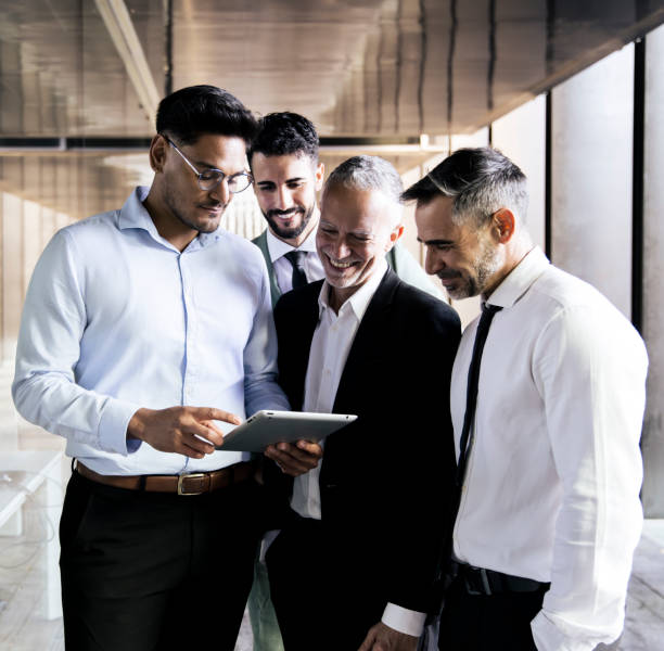 istock Diverse group of businessmen checking a tablet in an office corridor. Young executive holding a device in a meeting with senior colleagues standing at office. 1587804090