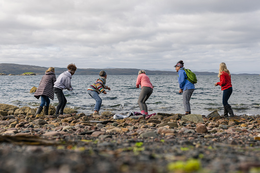 A rear view of a group of senior women who are enjoying a trip away to Loch Torridon in the Scottish Highlands. They are stood at the shore edge and skimming stones together and having fun together and seeing who can skim the furthest along the shore.