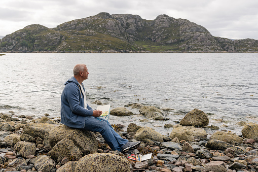 A wide angle side view of a man on his own sitting and journalling. He is sitting on a rock and documenting his trip along the Scottish coast in Loch Torrridon and having a mindful moment.