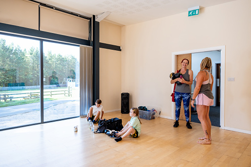 Two children sitting on the floor in a community hall where they're removing their shoes before their kickboxing class. A parent and instructor are standing close to them and bonding before the class.