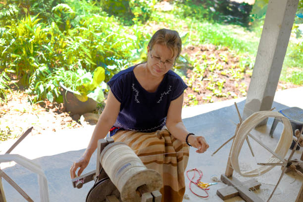 Mature thai woman is looming and spinning wool in On Tai Mature thai woman is looming and spinning wool in On Tai, Chiang Mai, San Kamphaeng district of Chiang Mai. Village Aree Pha Tor is home of weaving textile industry for classical thai clothings handmade with traditional looms in north Thailand. Woman is sitting outside of a house and is working with wool. In background are plants true thailand classic stock pictures, royalty-free photos & images