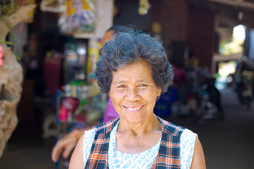 Toothy happy smiling  senior thai woman portrait outside of local rural store near On Tai, Chiang Mai, San Kamphaeng district. On Tai area disrtricts are local art and craft centers in  north Thailand. Authentic village life scene.  Woman has short curly hair and is looking and smiling at camera. In background is open entrance area