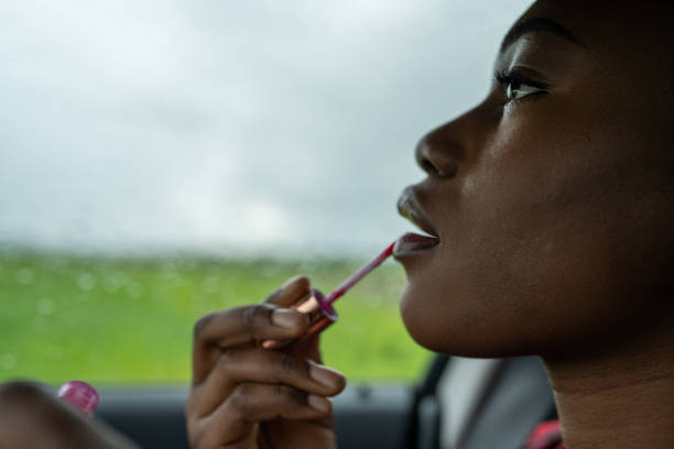 Black woman applying lip gloss in the car, it's a rainy day outdoors.