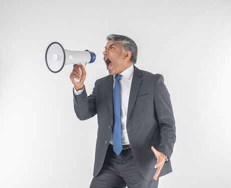Angry senior male professional dressed in suit screaming into megaphone while standing against white background