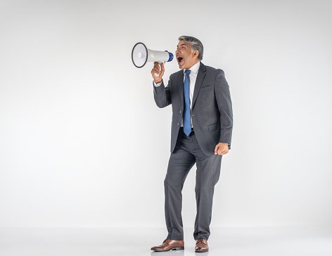 Angry senior male professional dressed in suit screaming into megaphone while standing against white background