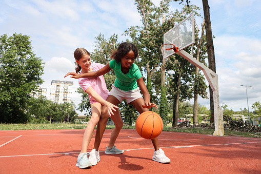 A multi-ethnic group of elementary age children are playing together at the gym during recess. One girl is holding a basketball and is smiling while looking at the camera.